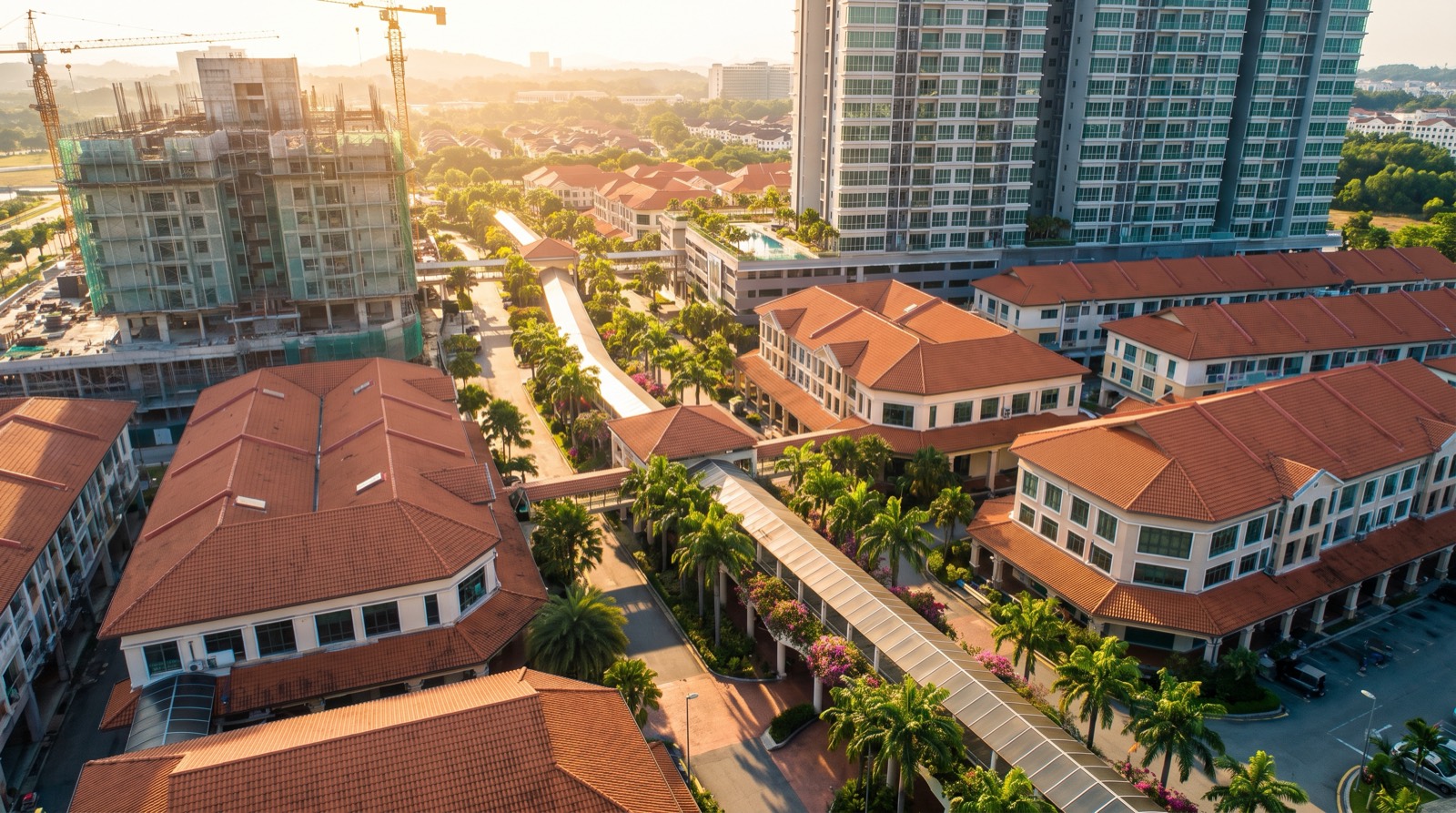 An aerial view at golden hour over a Malaysian new township, showing red-tiled terraced houses, a covered elevated walkway running through landscaped palms, a high-rise tower block and a building under construction with a tower crane