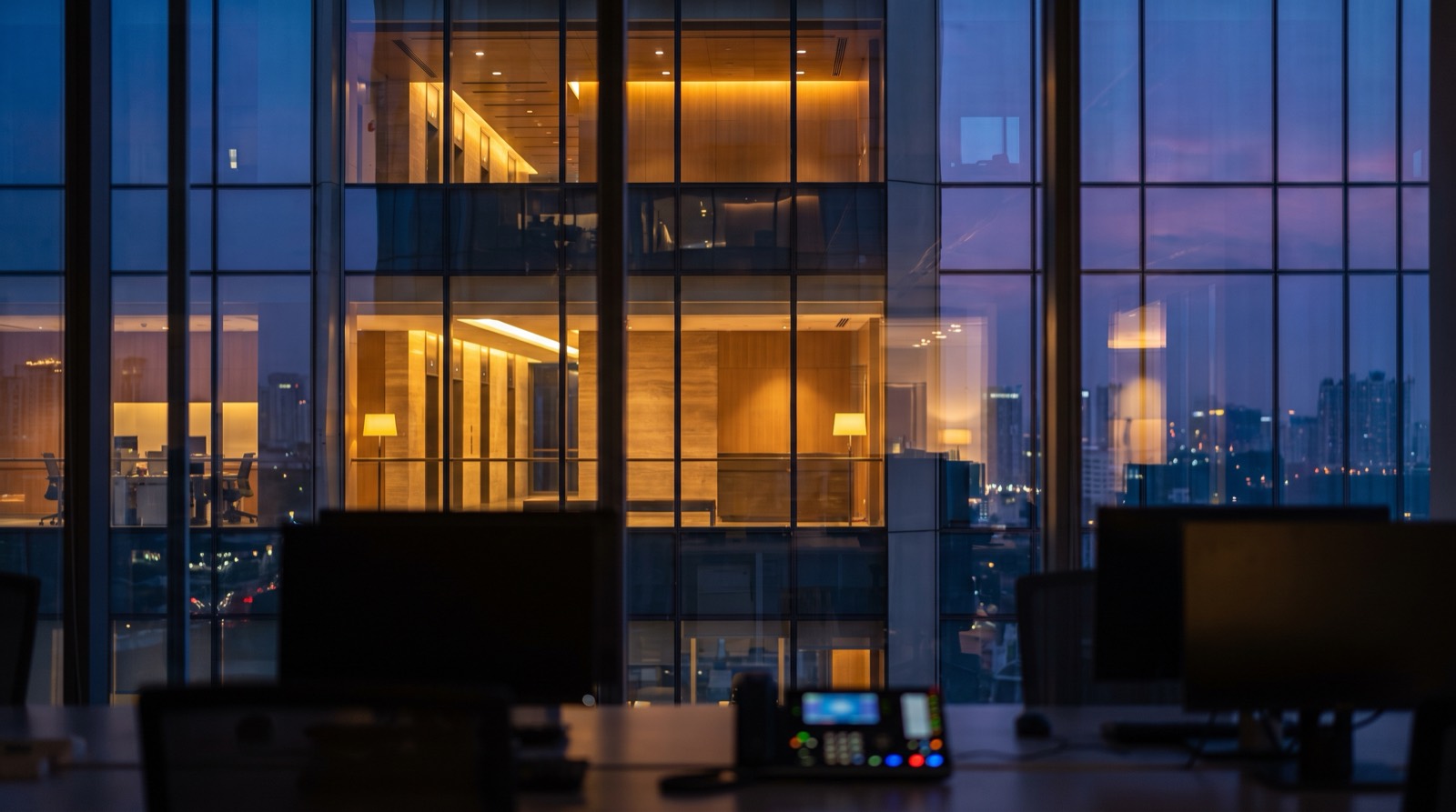 A darkened building operations desk at night, seen from behind: monitor backs and a lit control console in the foreground, looking out through a floor-to-ceiling glass curtain wall at a multi-storey office tower where several floors are lit and others are dark, with a city skyline beyond. The frame captures the actual subject of this post — not a device in a room, but one operator watching the state of an entire building at once.