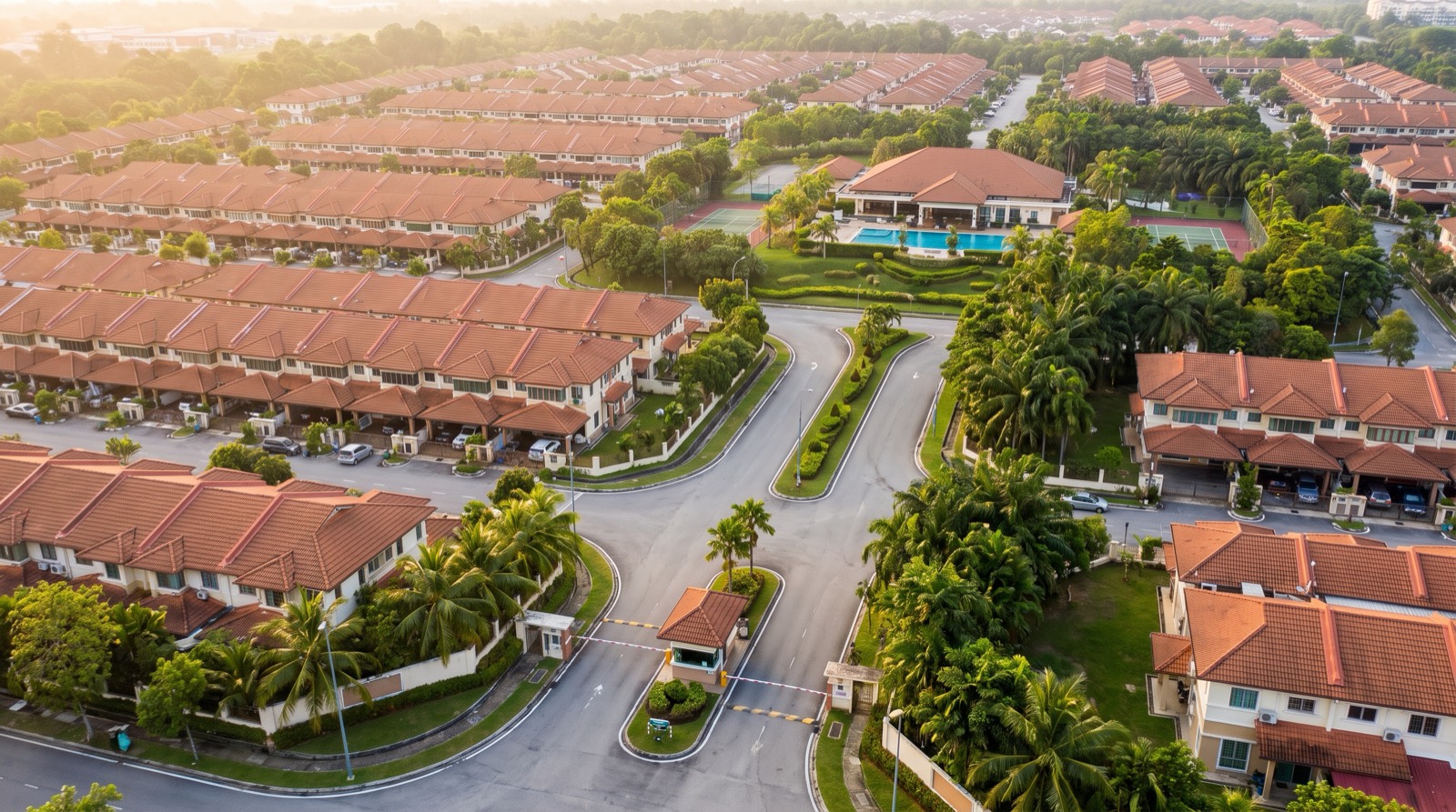 Aerial view of a large gated landed housing scheme at golden hour — rows of two-storey terrace houses with red-tiled roofs lining a curving internal road, a guard house and boom barrier at the entrance in the foreground, and a clubhouse with a swimming pool and tennis courts behind it, showing how many separate shared systems one township has to hand over at once