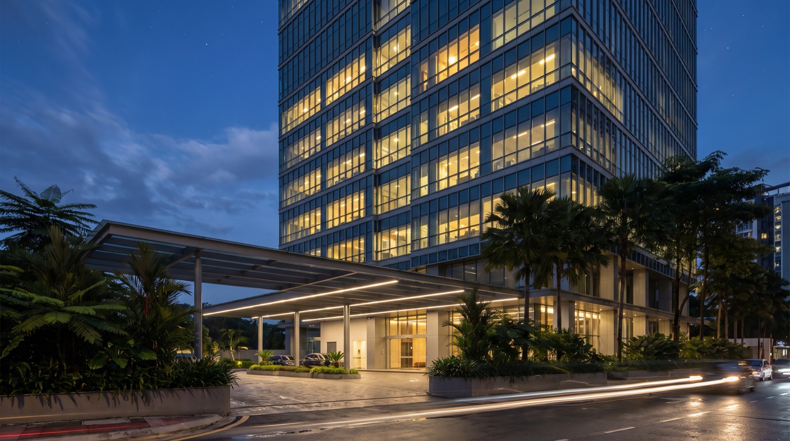 A modern commercial office tower at blue hour — a glazed multi-storey building with a scattered pattern of lit and unlit floors, a wide steel-and-glass entrance canopy lit by linear strip lights, landscaped planters with palms and tree ferns, cars parked beneath the canopy, and a wet road in the foreground with light trails from passing traffic — a whole building rather than one room, because a commercial quote is never assembled room by room
